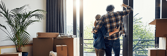 Photo of a man and woman embracing in front of the window of their new home with moving boxes