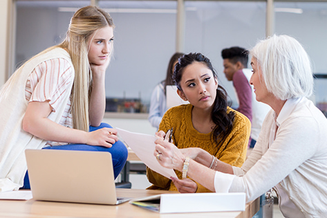 Two female students sitting at a table chatting with a teacher