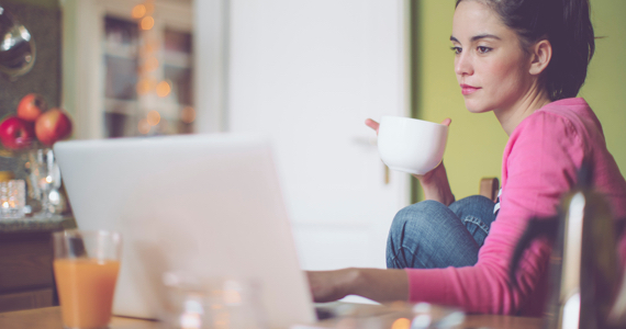 Young relaxed woman drinks her coffee in her kitchen while looking at her computer