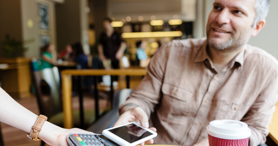 Man in a coffee shop taps his phone against a payment terminal