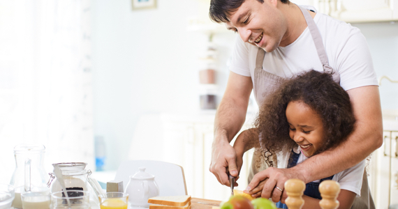 Man in a kitchen helps his little girl cut a slice of bread