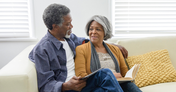 Elderly man and woman sitting on a couch smile while looking at each other
