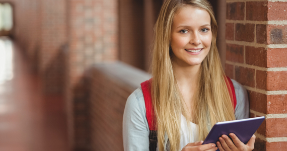 Young female student in a college hallway is holding a tablet while smiling