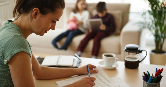 Woman works at her desk while her two children are reading on a couch