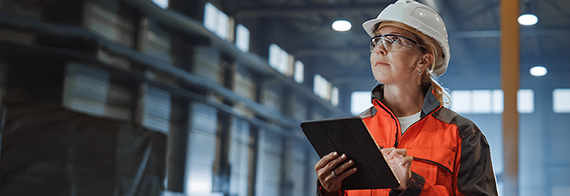 Photo of a woman wearing a construction helmet and holding a tablet in a large building 