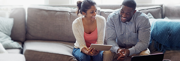 Couple smiling, sitting on a sofa, looking at a tablet