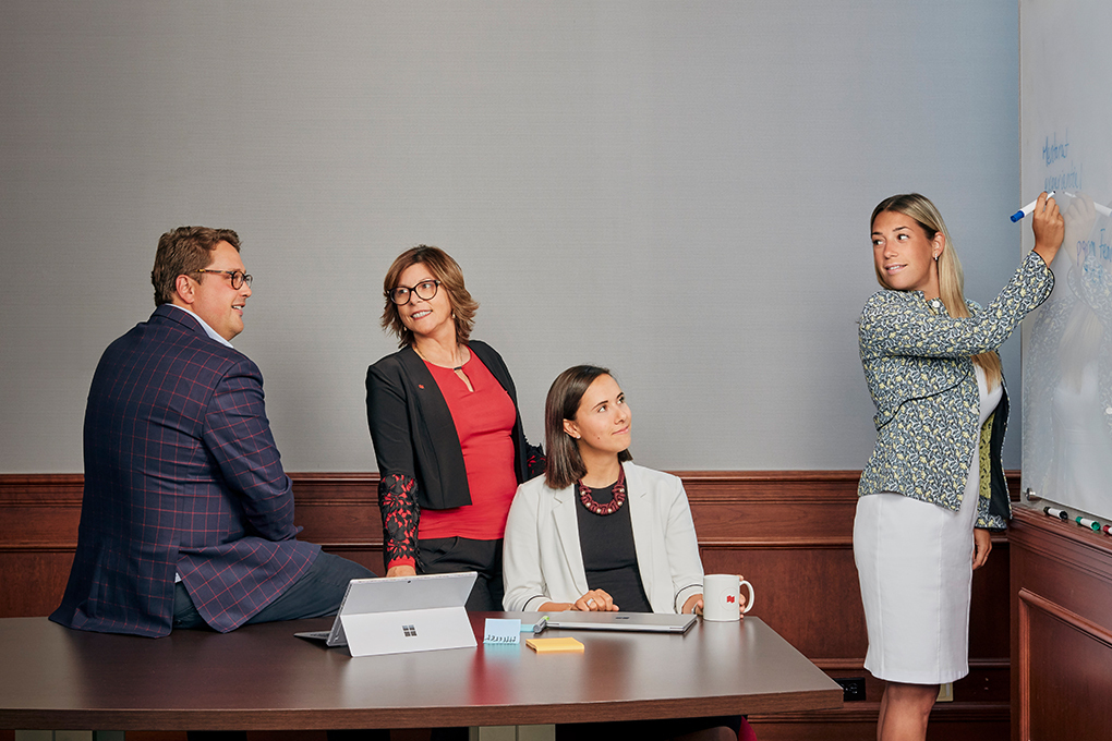 Photo of a woman writing on a blackboard in an office in front of a man and two women in professional clothing 