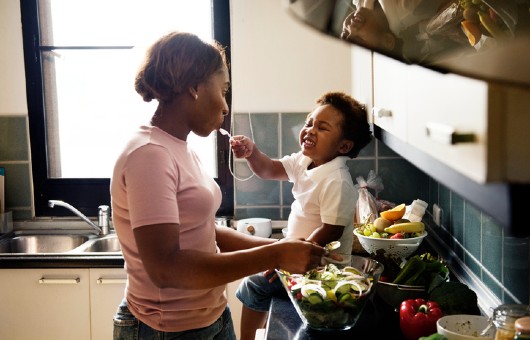 A kid feeding his mother while she’s cooking.