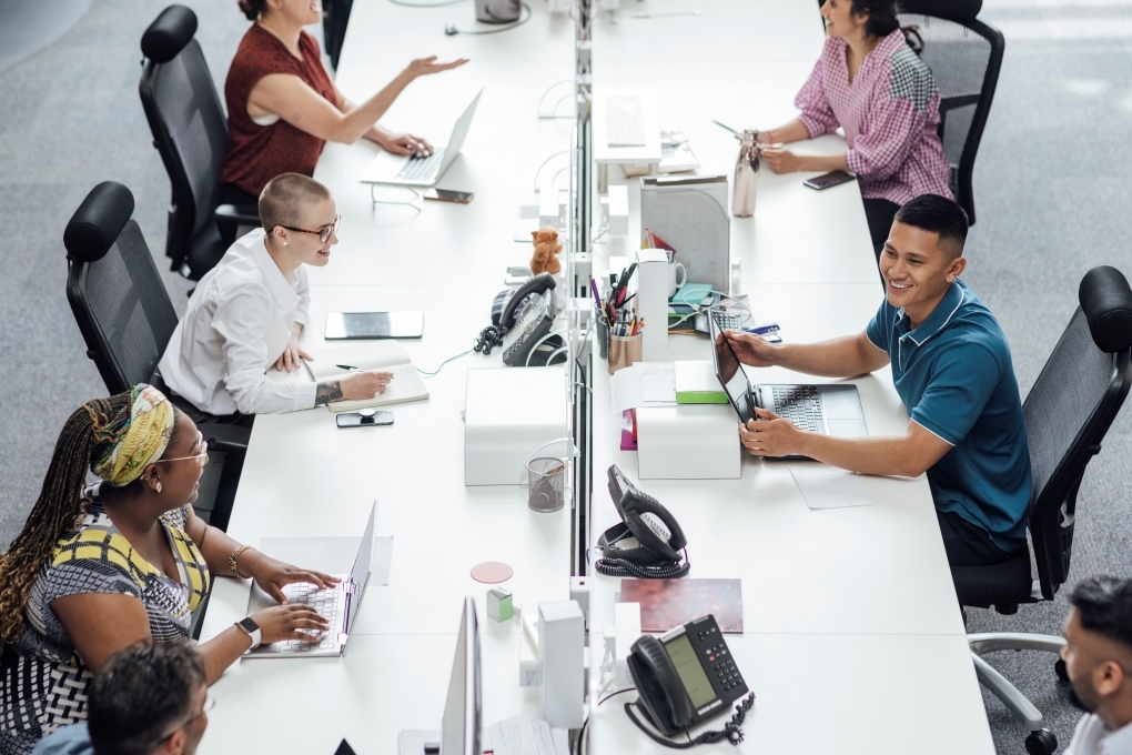Photo of a shared office with several young people working, chatting, and smiling while using laptops. Office supplies are scattered on the tables.