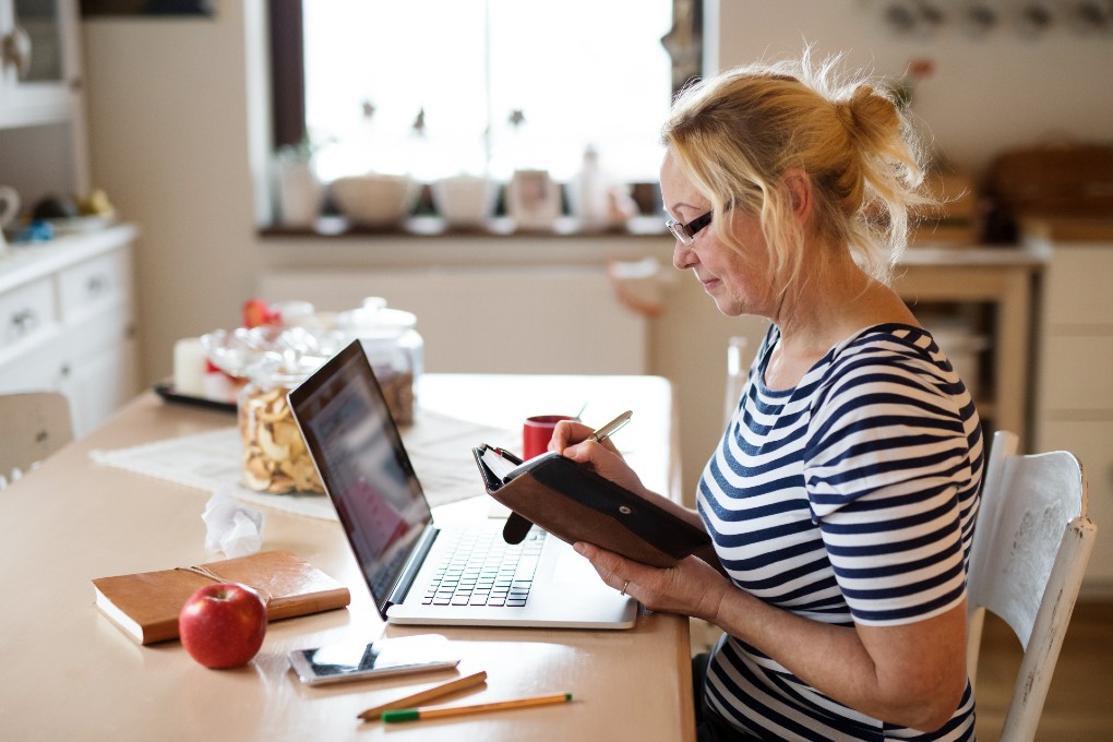 Image of a woman sitting at her kitchen table takes a note in a notebook.