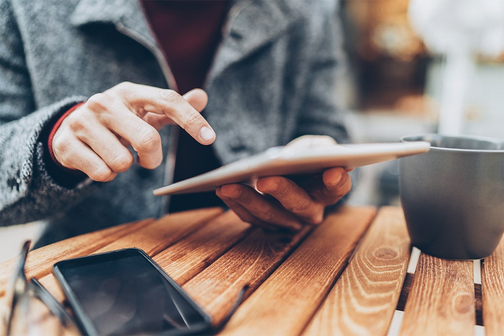 Image of a man's hands using a tablet on a table with a cup and a cell phone.