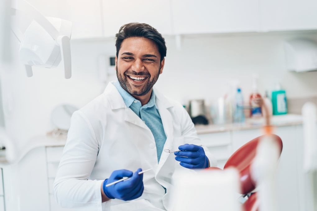 A dentist in a white lab coat, holding a dental tool is smiling in his office.