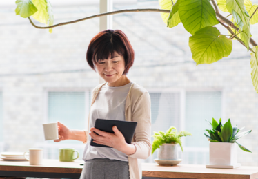 Photo of a woman in front of a window consulting her tablet with a cup of coffee in her hand 