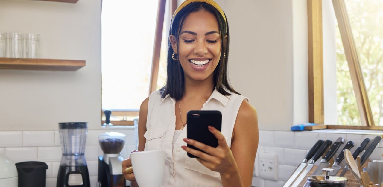 Photo of a smiling woman standing in her kitchen holding a cup of coffee and her phone