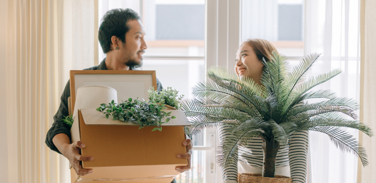 Photo of a couple carrying a box and a potted plant into their new home 